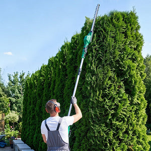 A man in gray overalls and a white t-shirt uses a long-reach hedge trimmer to cut down a tall green evergreen hedge in a sunny backyard garden.